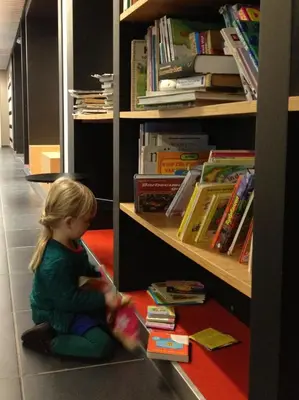 a little girl sitting on the floor in a library at Hostel De Veurs in Sint-Martens-Voeren