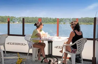 two women sitting at a table by the water at Camping De Binnenvaart Superior Chalet in Houthalen-Helchteren